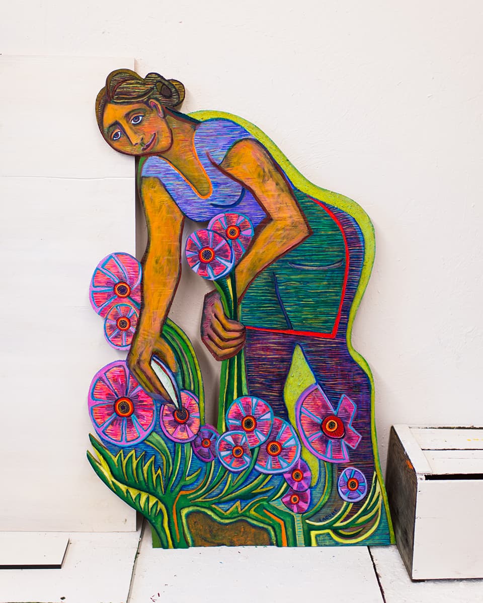 Photo by Robert Jaffe of artist Betty LaDuke’s 6 feet tall carved and painted wood panel of a flower farmer cutting colorful flowers with a harvest tool, on the fields of Le Mera Gardens.