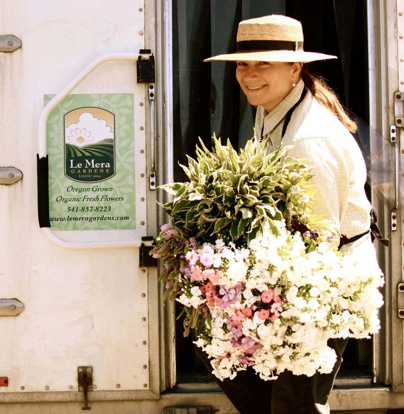 Flower farmer and Slow Flowers advocate Joan Ewer Thorndike wearing a sun hat on the steps of a flower truck holding bunches of bi-colored dogwood foliage, physostegia and phlox freshly harvested from the fields of Le Mera Gardens.  Le Mera Gardens logo and tagline on a sticker adhered to the back wall of the flower truck.